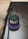 New Era 59Fifty HWC '93 Wordmark Milwaukee Bucks Fitted Hat In Green & Purple - Top View Lifestyle Photo On Ground